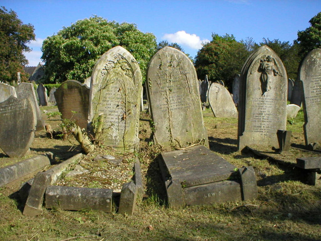 A cluster of old graves a gravestones in a cemetery with trees in the background and a blue sky