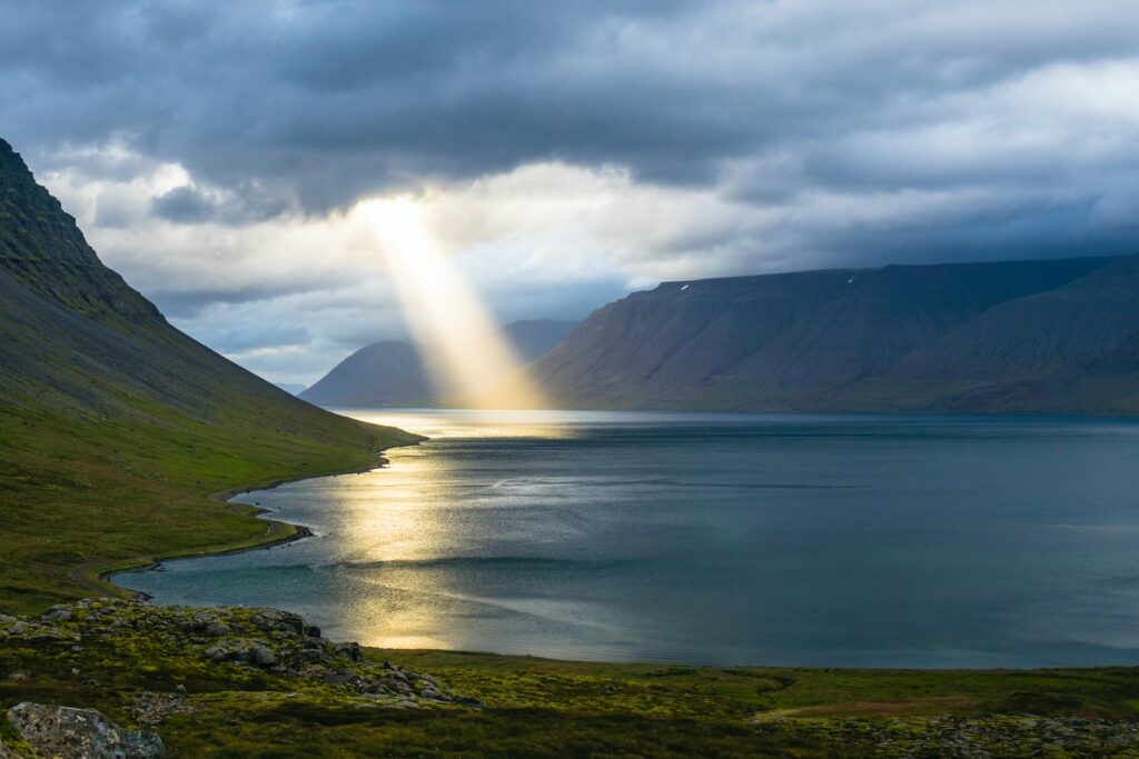 A photo of a wide calm waterway between two low lying mountains with a large beam of sunlight, in a heavily clouded sky, laying across the water in the distance