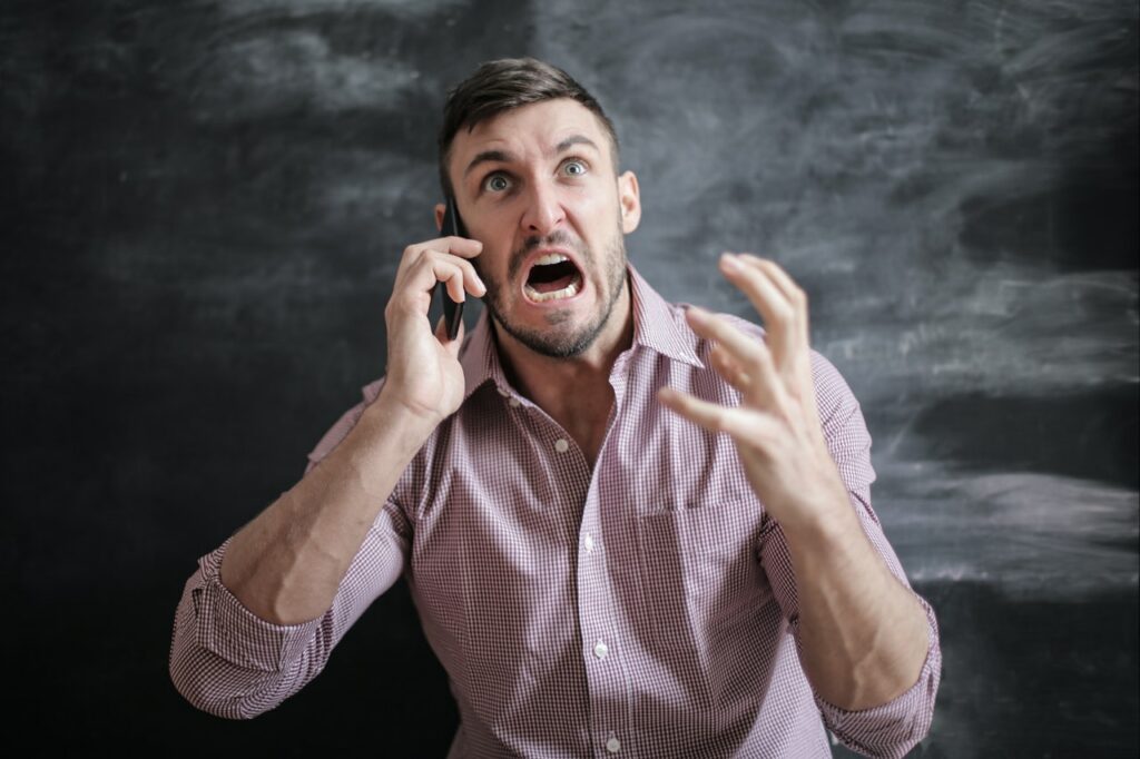 A man in a pale pink buttoned shirt talking on a phone with arms partially upraised in an animated manner while angry thoughts are being expressed on his face.