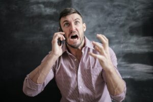 A photo of a Caucasian man in a pink button shirt with dark hair and an angry face looking upward while shaking his hand and holding a mobile/cell phone to his right ear