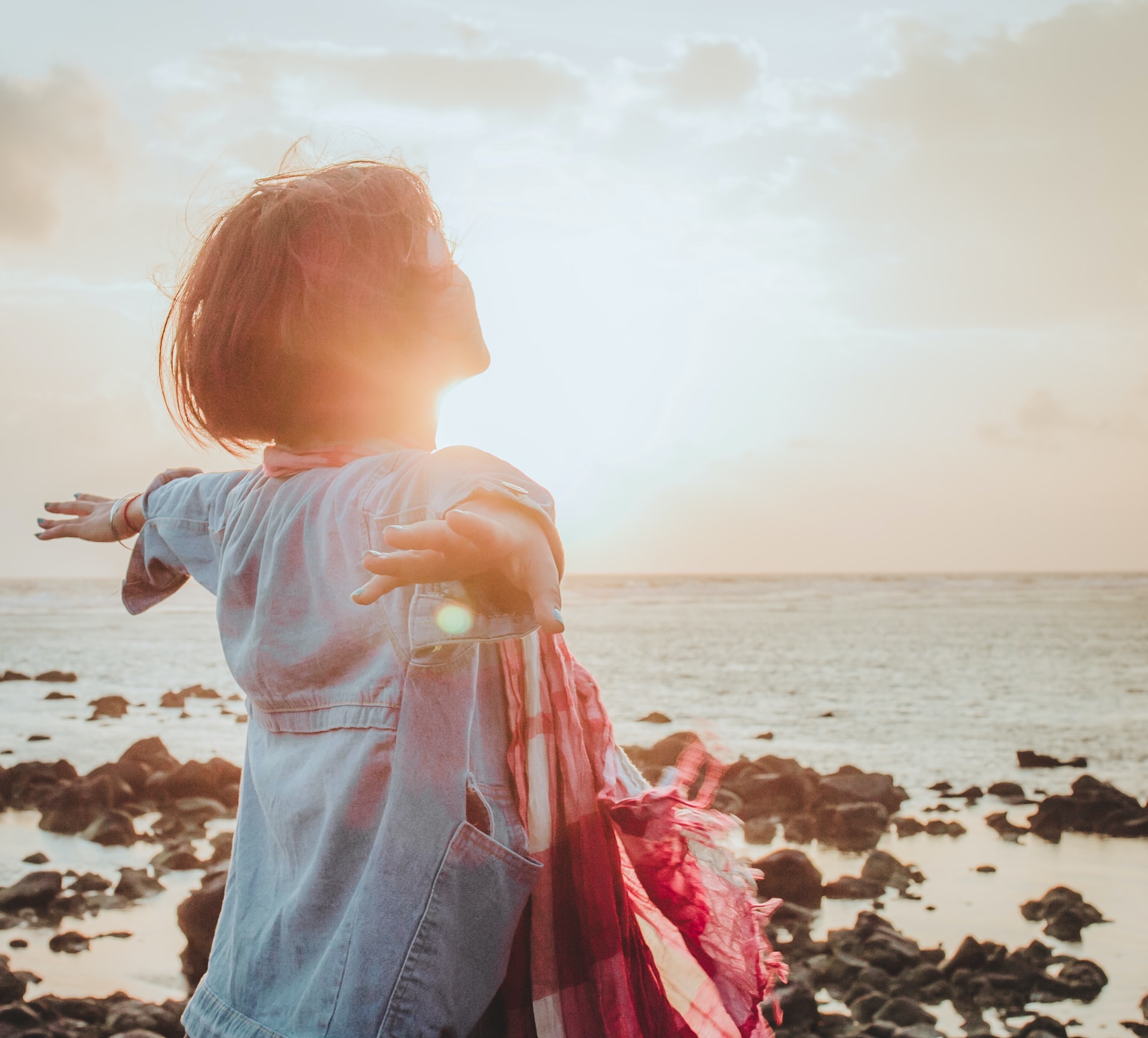 A woman standing at a rocky seashore facing towards the water with her head up and arms outward while wearing a light blue denim shirt and a red plaid scarf around her neck with a hazy sunlight sky in the background