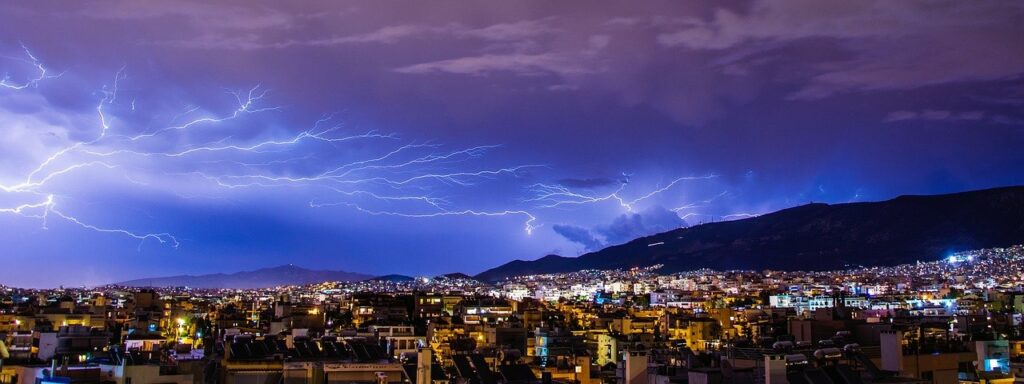 A large thunderstorm and much lightning illuminating the sky with a blue tint over a vast city in the evening with mountains in the distance