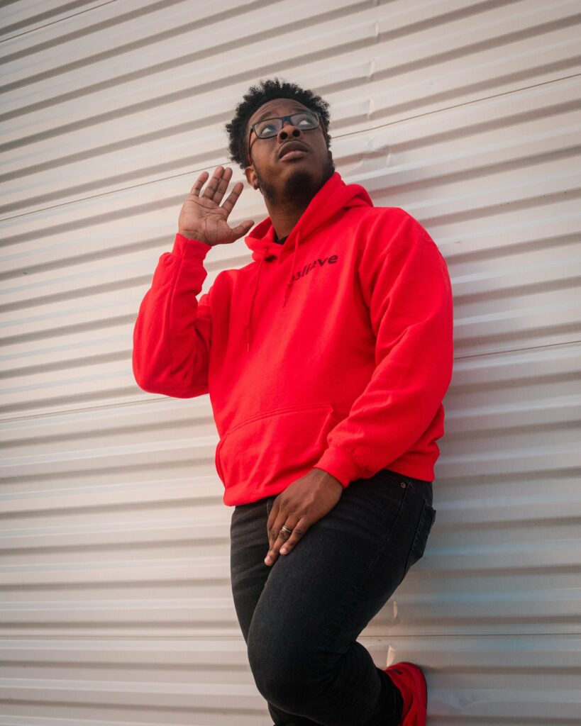An African-American man wearing a bright red hooded sweatshirt and black jeans leaning against a steel paneled wall looking upward toward his backside with his open right hand held up near his right ear