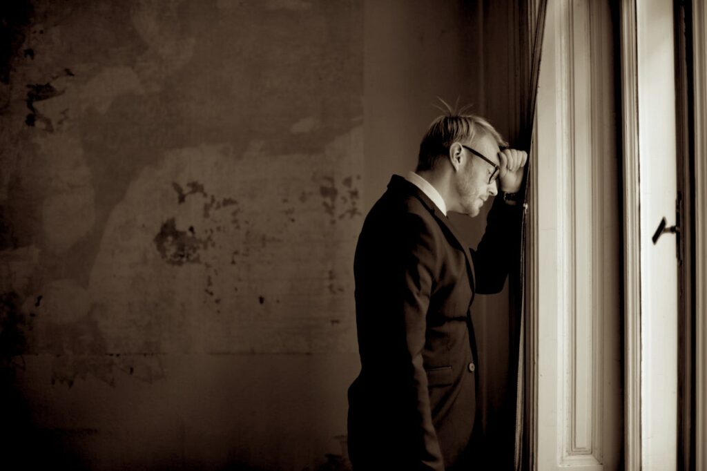 A black and white photo of a older Caucasian man wearing a suit and glasses while standing and leaning with his hand against a window frame looking slightly downward with his eyes closed in an old room
