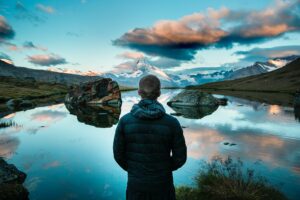 The backside of a young man in a dark winter coat facing a large still lake with mountains, blue sky, and clouds in the distance