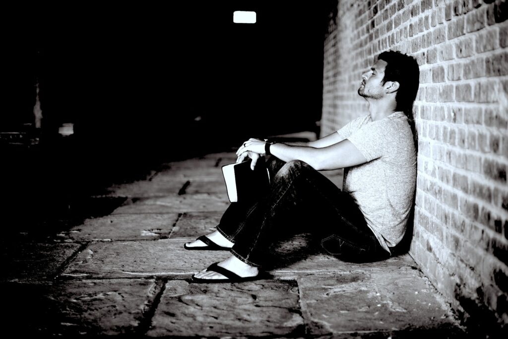 A Caucasian man seated on a stone tiled ground against a brick wall in an old building with legs arched, hands holding a Bible and eyes closed in prayer