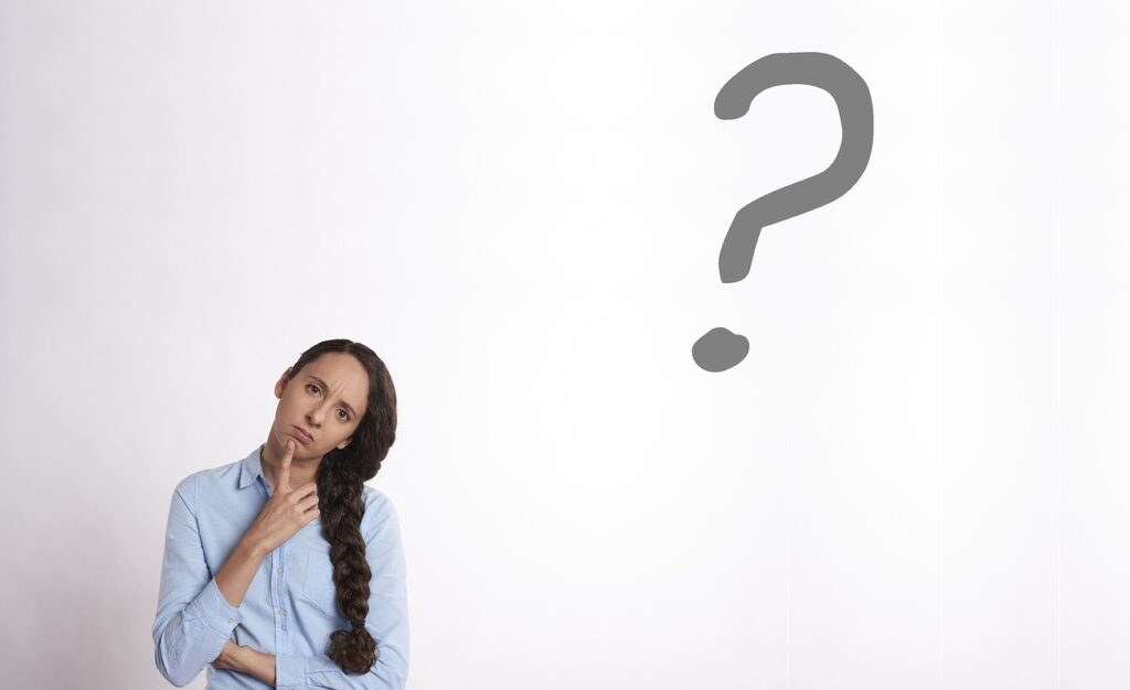 A young woman in the lower left corner of the picture wearing a light blue long sleeve shirt and black braided hair with her head cocked to the right and her right index finger held up to her chin with a large grey question mark in the upper right corner of the picture