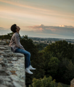 A photo of a young Hispanic man sitting on a long concrete wall on a hillside looking upward with a pale clouded sky in the background