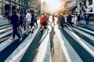 A photo of a large group of people walking across a city street on a crosswalk with the sun streaming from behind