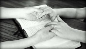 A black and white photo of two pairs of Caucasian hands being held while resting across an open Bible on a wooden table