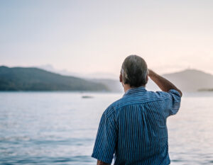 A photo of the backside of an older man in a blue striped short sleeved shirt holding his right hand against his forehead while looking across a large body of water with small mountains in the distance