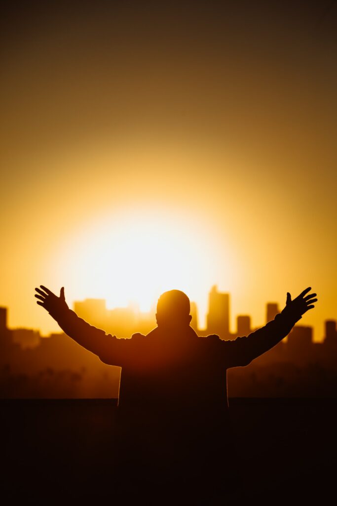 A silhouette of a man with his arms raised while facing an urban skyline with a bright yellow sun in the background