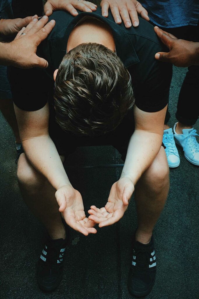 A Caucasian man sitting with his head down and arms leaning outward resting on his knees with several hands resting on his back