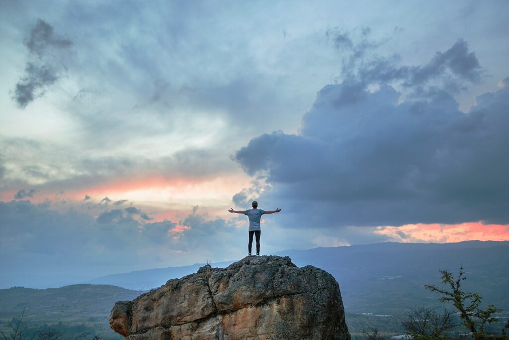 A person standing on a large tall rock formation with their hands upraised while facing a blue tinted cloudy sky with reddish tints of a sunset peeking through and mountains in the distance while recognizing the love of God.