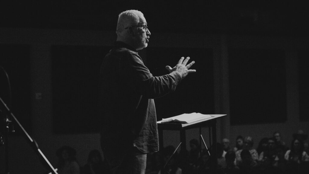 A black and white photo of the side view of an older man with white hair and a suit standing at a podium with hands partially raised before a large group of people