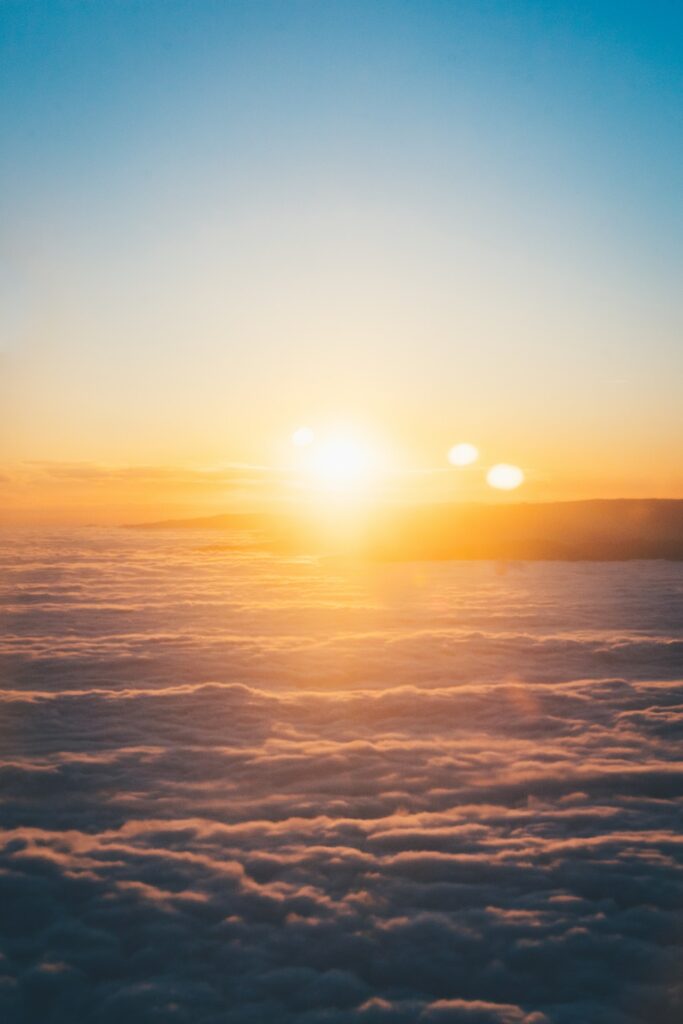 A photo of a bright yellow-white sun above the clouds resting on top of a small mountain against a light blue sky