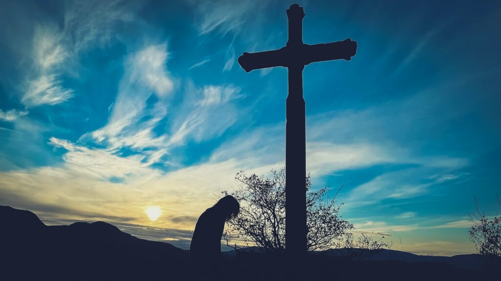 A photo of the silhouette of a large cross and a young woman kneeling at the base with a bush and some mountains in the background against a blue sky and a setting sun lightly obscured by cirrus clouds