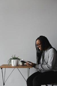 A smiling African-American woman with glasses and long hair seated in a chair wearing a grey long sleeved shirt and black pants while facing a small table with a small plant in a white ceramic pot against a grey wall