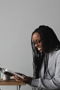 A smiling African-American woman with glasses and long hair seated in a chair wearing a grey long sleeved shirt and black pants while facing a small table with a small plant in a white ceramic pot against a grey wall