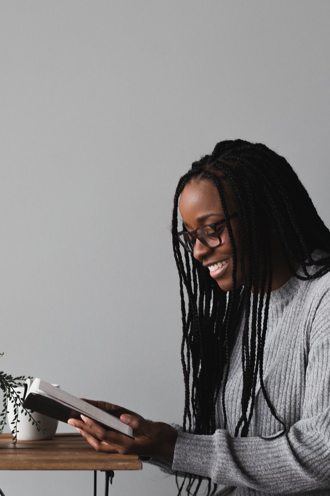 A smiling African-American woman with glasses and long hair seated in a chair wearing a grey long sleeved shirt and black pants while facing a small table with a small plant in a white ceramic pot against a grey wall