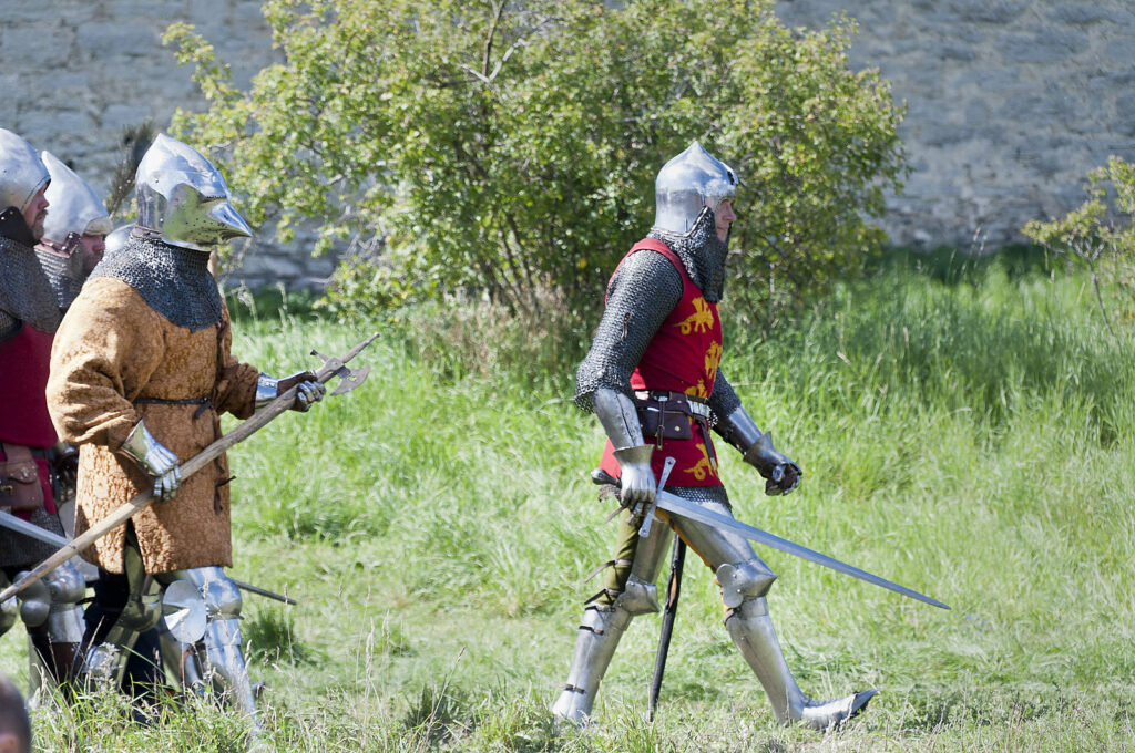 A photo of several Medieval dressed soldiers in full armor in reenactment of the battle of Wisby of 1361 with a large green shrub and a hillside in the background