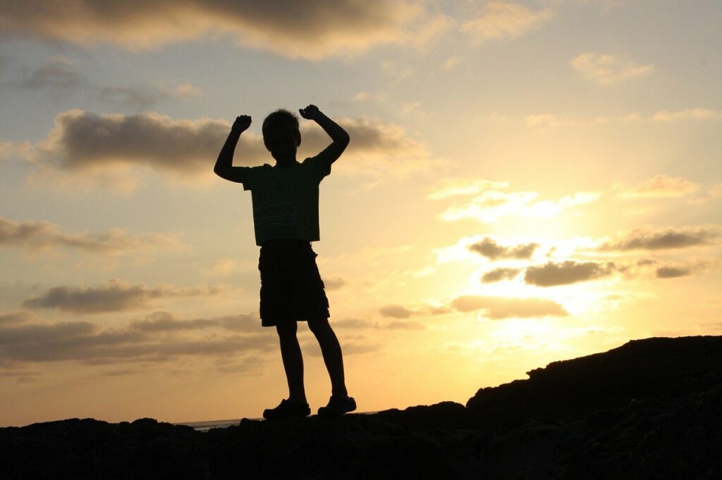 A silhouette of a young boy in shorts and a t-shirt standing on a small hill with his arms raised in the air and a setting sun among assorted clouds in the background