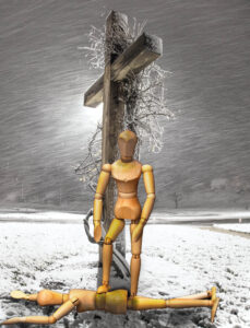 A composite photo of a wooden artist's model standing with its foot against another artist's model laying prostrate on a snowy ground with a large wooden backlit cross immediately behind them against a dark sky and blowing snow