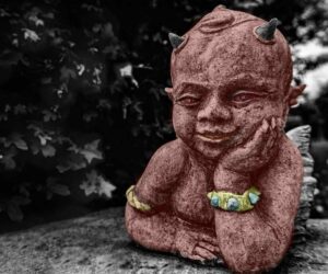 A modified photo of a young devil sculpture leaning against a flat stone surface while resting his head against his left hand with right hand folded against his chest and a low hanging maple branch in the background