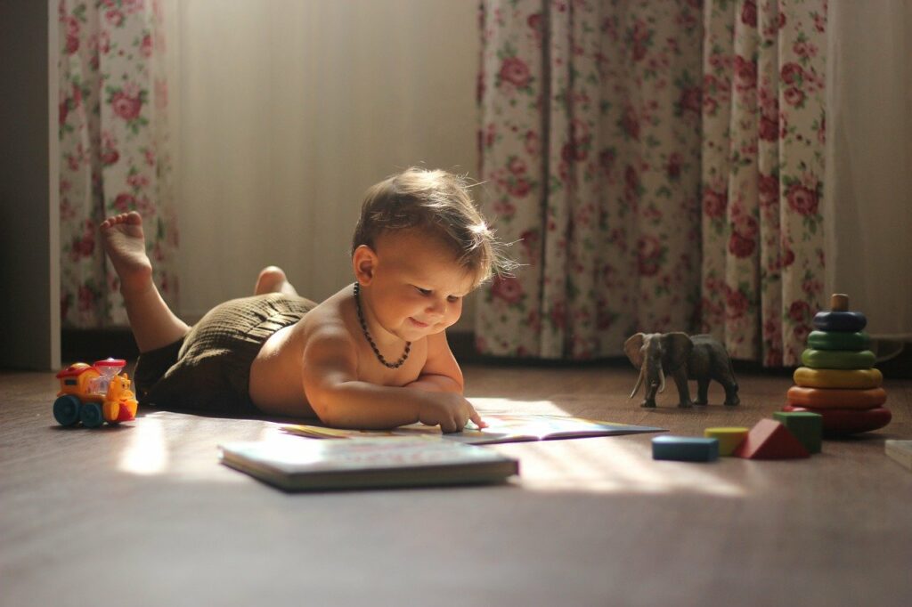 A young Caucasian boy wearing green shorts and lying on his stomach reading with sunlight streaming over him in a room with several toys and flower print curtains in the background