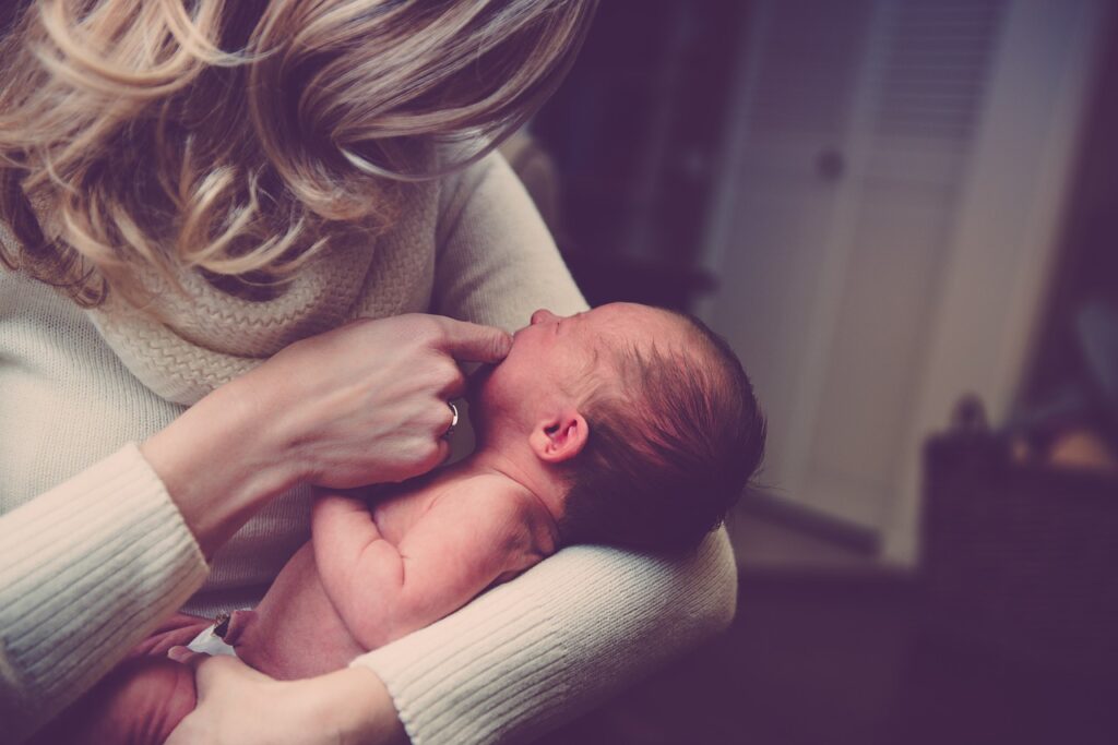 A woman with long blond hair and a white turtleneck sweater holding a newborn infant in her arm while touching its mouth with her other hand