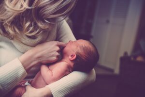 A woman with long blond hair and a white turtleneck sweater holding a newborn infant in her arm while touching its mouth with her other hand