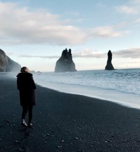 A woman with dark hair walking along a shoreline comprised of black sand and stones while looking toward a large body of water on the right with assorted eroded rocky pillars in the distance