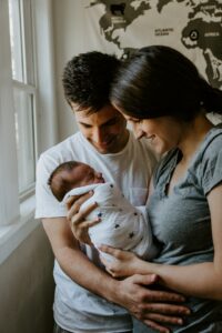 A young couple holding and looking at a new born child wrapped in a blanket while standing in front of a window