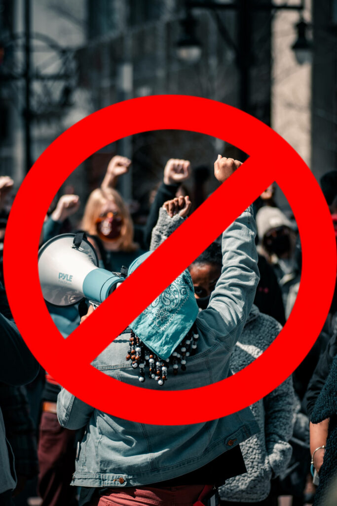 A modified image of a red circle with a diagonal slash overlayed on a photograph of several people in a city holding their fists in the air including the backside of one wearing a light blue bandana on her head while holding a megaphone in her left hand