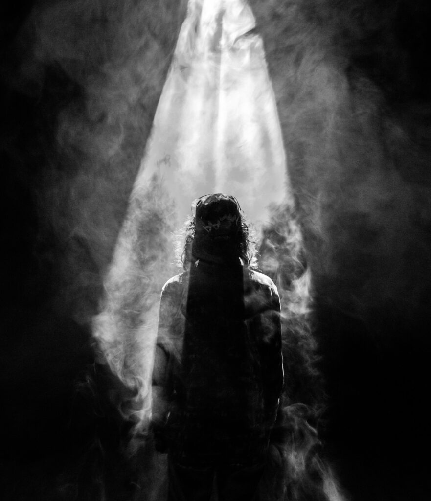 A black and white photo of the back of a standing man surrounded in fog/smoke with a beam of light shining on him from above