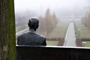 A photo of a close up of the backside of a seated male statue overlooking from an elevated position, a paved pathway with single columns of trees nearby on each side and a foggy background mostly obscuring a row of homes and trees
