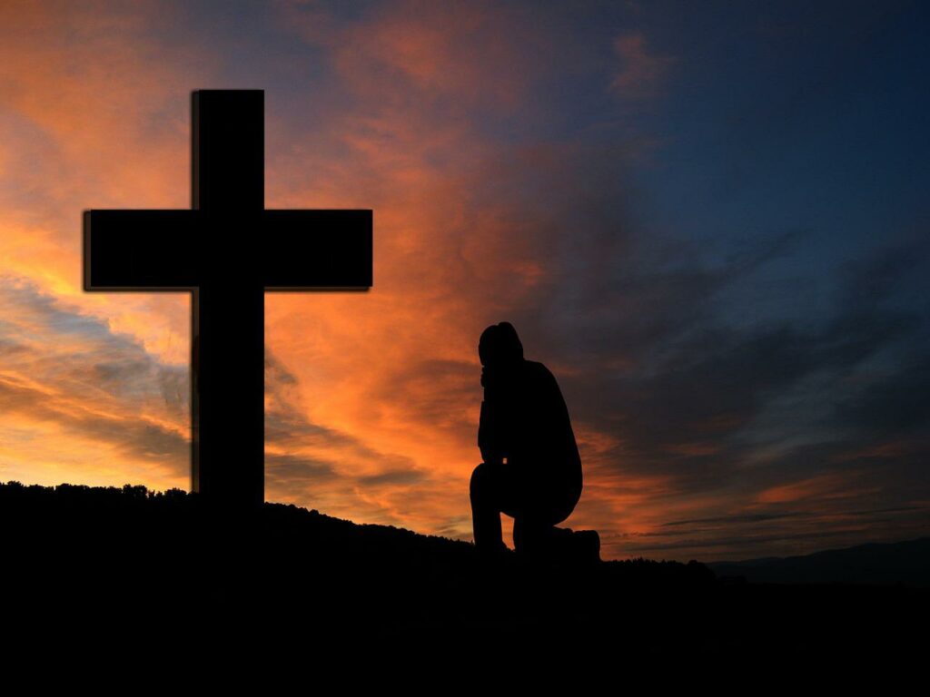 A silhouette of a man kneeling in front of a short cross with clouds colored with various shades of orange from a setting sun against a dark blue sky in the background