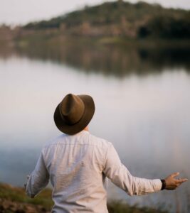 A photo of the backside of a man wearing an olive green hat and a long sleeved white buttoned shirt with hands partially upraised while facing a large still lake and a hill in the distance