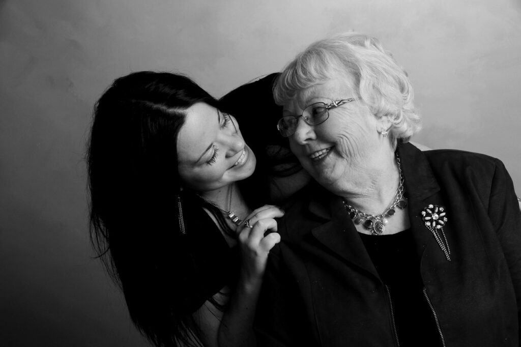 A black and white photo of a elderly Caucasian woman with white hair and a dark blouse smiling while looking back at a younger Caucasian woman with black hair leaning over and smiling at the elderly woman