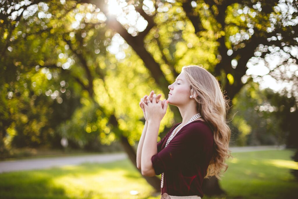 A photo of a Caucasian woman with blond hair standing in a park with trees and sun in the background while holding her hands together near her face and her eyes closed