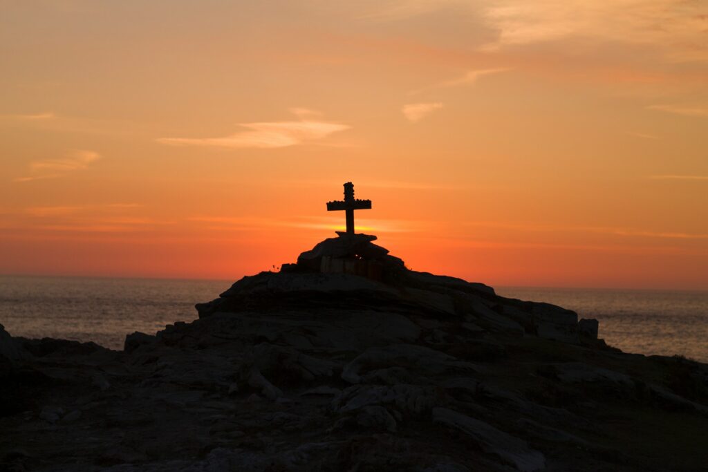 A silhouette of a cross on a small rocky hill with a orange sunset in the background