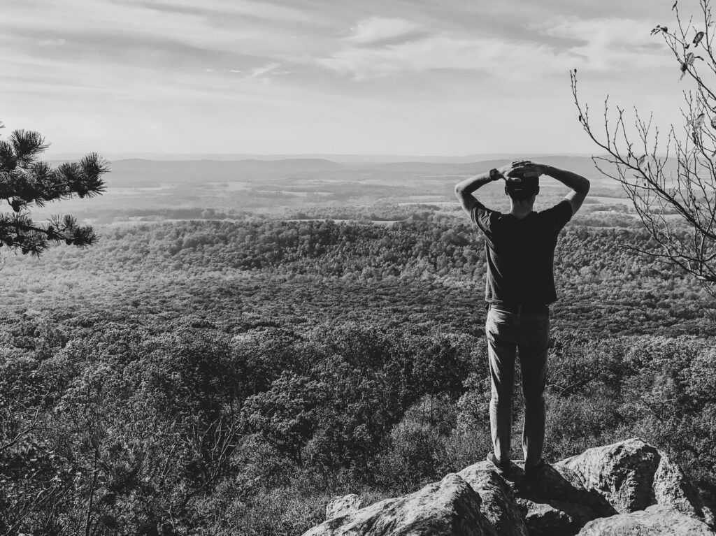 A man looking out from a mountain ledge at the peaceful valley below.
