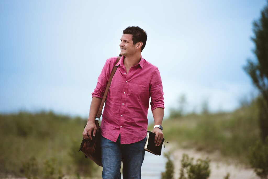 A smiling man wearing a red buttond shirt and blu jeans walking on a boardwalk among dunes while carrying a bag an a Bible showing the importance of being confined to God and His Word rather meddling in other's affairs