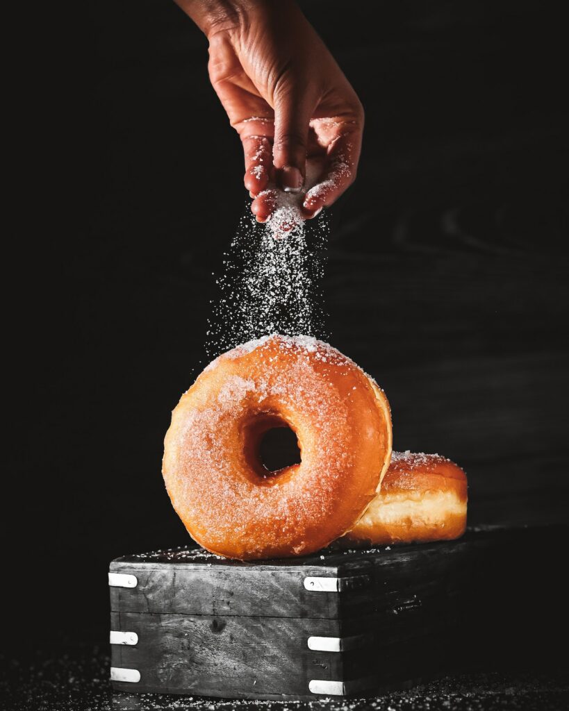 A pair of glazed donuts resting on a black box among a black background with sugar being sprinkled from above and coating them like the truth of sin being sugarcoated.