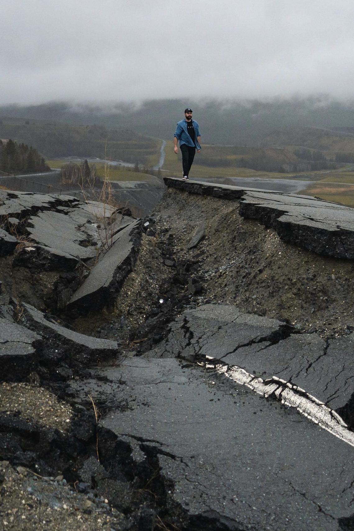 A man with a blue jacket and dark pants walking alongside a collapsed roadway from an earthquake with mountains and heavy fog in the distant background