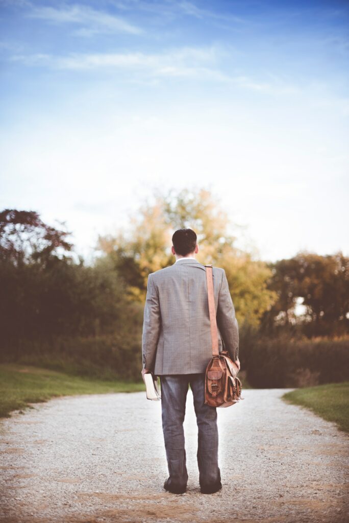 A dark haired man wearing a grey suit jacket and casual pants carrying a leather book bag on his right shoulder and holding a Bible in his left while depicting a righteous demeanor.