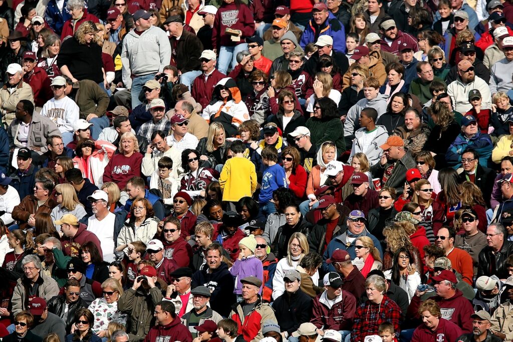 A large assorted crowd of people mostly seated, wearing moderately warm clothing on a sunny afternoon symbolizing how the grace of God is available to everyone