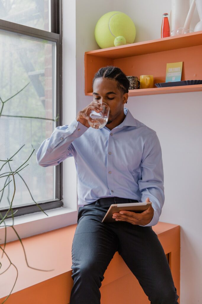Man taking a moment to drink from a glass of water while holding a tablet and sitting on a counter in a room.