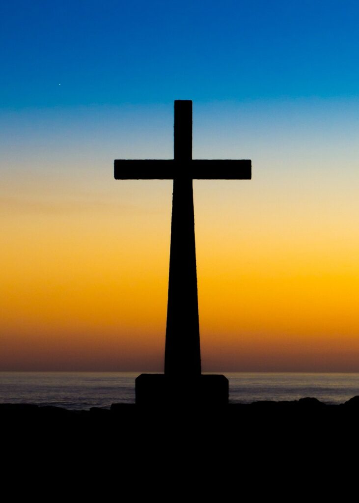 A silhouette of the cross of Christ on the shore of a large body of water against a gradient sunset background