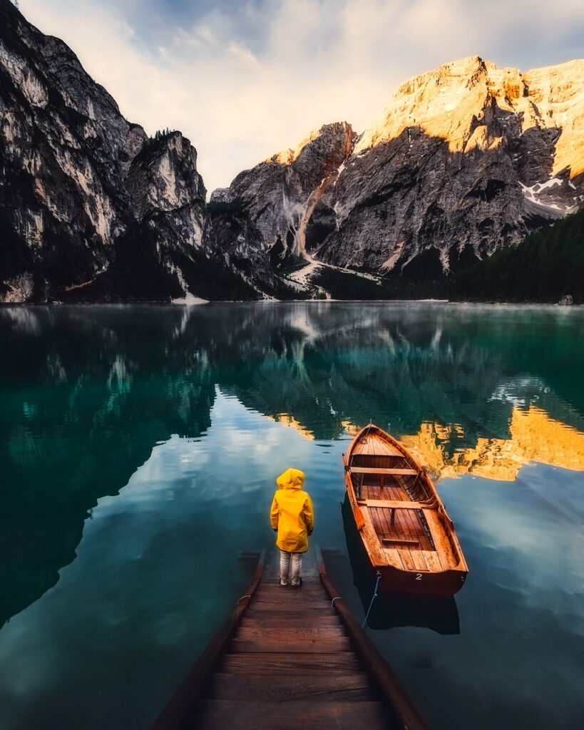 A person in a yellow coat standing on a small dock partially underwater next to a moored wooden boat while looking across a calm lake with mountains in the background. Demonstrating the need to go when Jesus compels us.